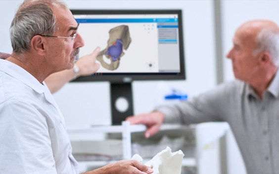 Doctor holding a 3D-printed anatomical model and pointing to a screen including the digital design of a personalized hip implant while talking to a patient
