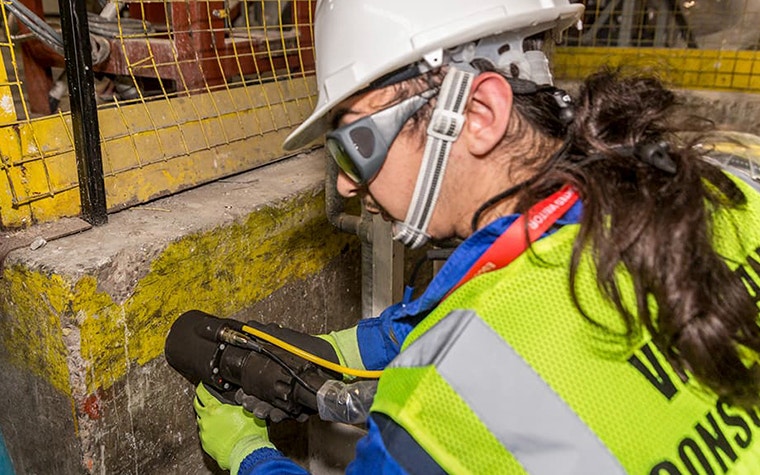 A Viridian consultant wearing a fluorescent vest, helmet, goggles, and gloves takes a reading from a wall using the sampling head 