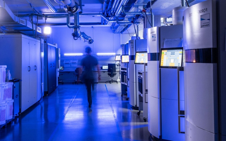 A blurred person walking through a row of 3D printers in a Materialise production facility