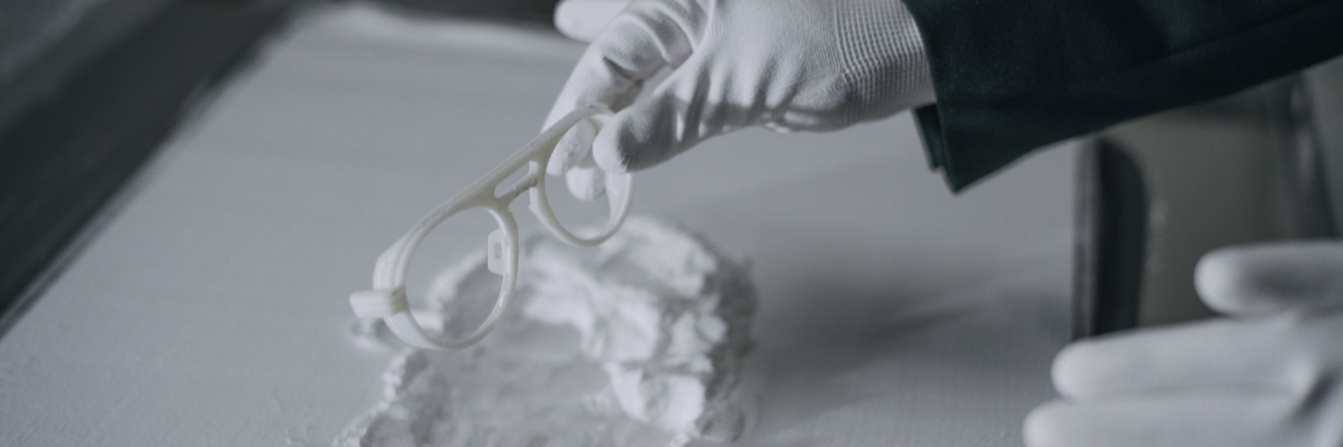 Mani guantate rimuovono le montature degli occhiali Odette Lunettes stampate in 3D da una letto di polvere