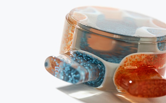 Detalle de una parte transparente impresa en 3D con partículas naranjas y azules en su interior, de lado
