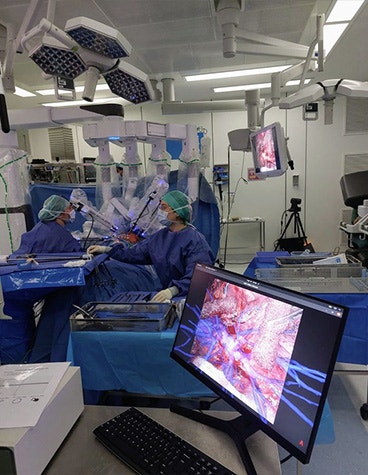 An operating room with three people in scrubs surrounding the surgical table. Screens hanging from the ceiling are on either side of the table, displaying a view of the patient's anatomy. Another person in scrubs stands back from the operating table, next to a table of surgical equipment.