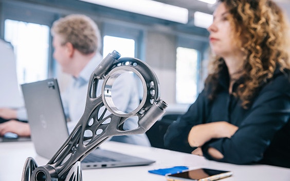 A metal 3D-printed part on a desk with two trainees sitting behind it on their laptops