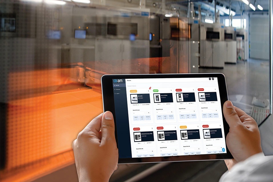 Persona che sorregge un tablet con la piattaforma software CO-AM di fronte a una stampante 3D