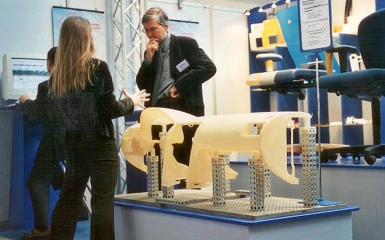 At the EuroMold event, two people stand in discussion beside the first demo part printed by a Mammoth printer. 