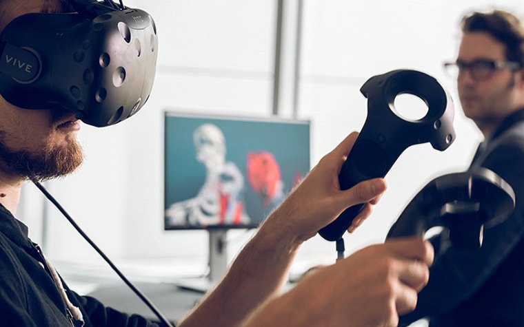In the foreground, a man wearing virtual reality goggles and holding controllers; in the background, a man sitting at a computer screen 