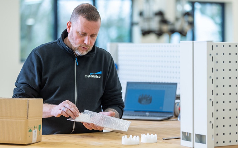 A Materialise team member putting a 3D-printed part into bubble wrap for shipment next to a packing box and a laptop with the design of the part