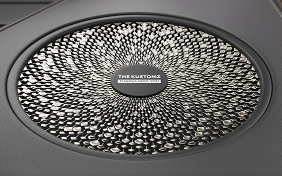 A speaker grill is shown mounted on a leather-look panel for a luxury car interior. The grill is personalized with a band name in the centre and 3D-printed in metal, with intricate diamond and bar shapes radiating into a circular pattern.