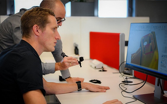 Two people comparing a 3D-printed part to a 3D design on a computer screen