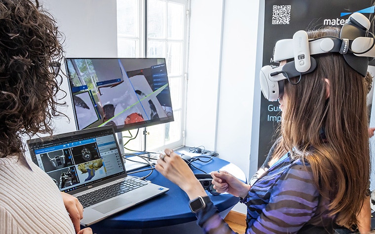 One woman wearing an XR headset at an event, holding out her hands to interact with a virtual model, while another watches on computer screens.