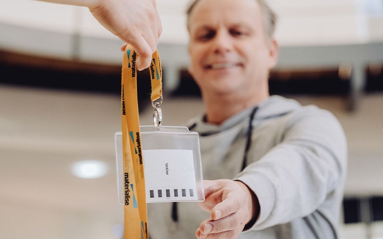 An image of a man holding out his hand reaching for a materialise visitor's badge. The badge has a yellow lanyard with the Materialise company name and logo.