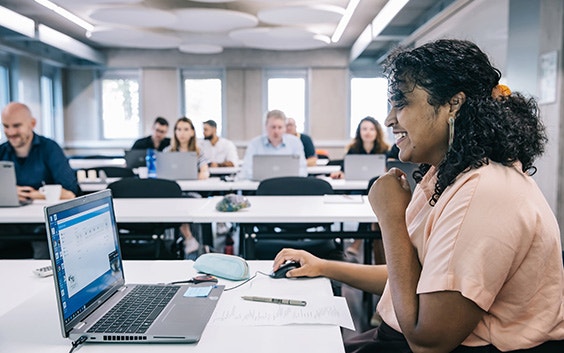 Woman sitting at a computer and smiling in front of a classroom of students on their laptops