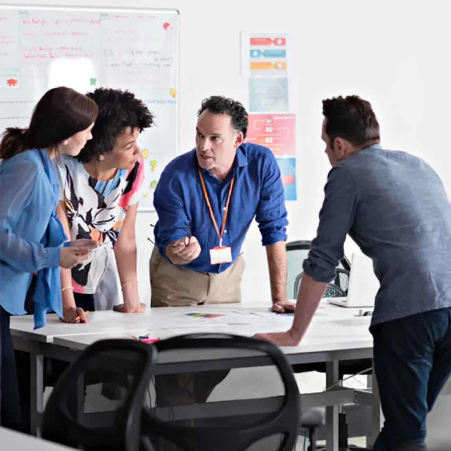 a man stands around a table with colleagues while he emphasizes his point by holding and pointing with his glasses. His coworkers feign interest.