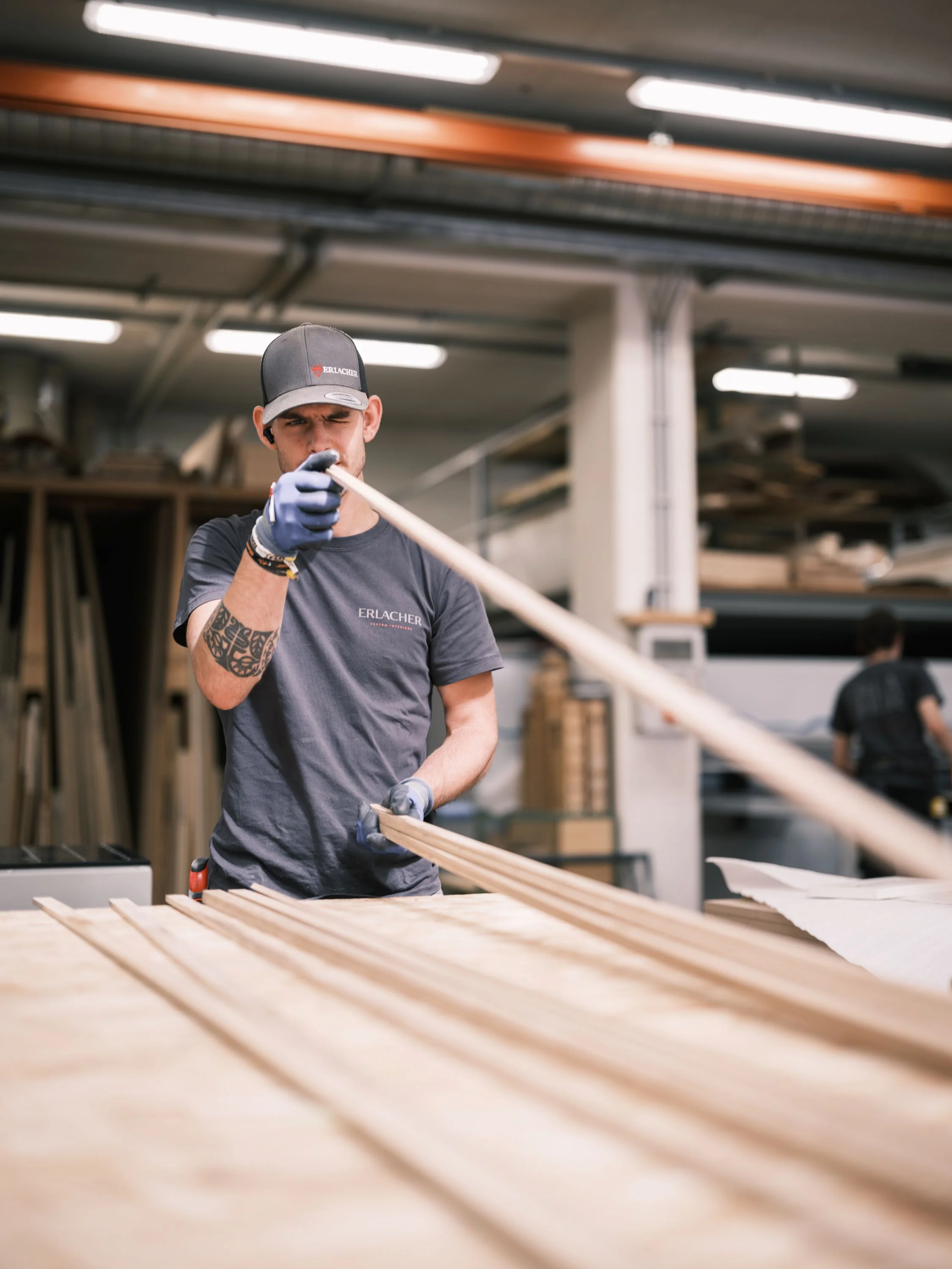 Craftsperson in a workshop wearing gloves and a cap, inspecting a long wooden strip on a workbench.