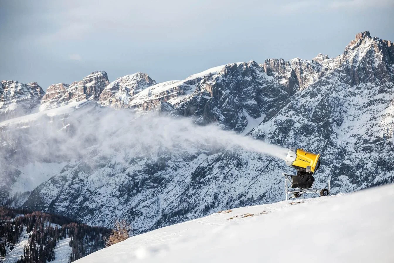 A snow cannon in action on a slope with mountains in the background