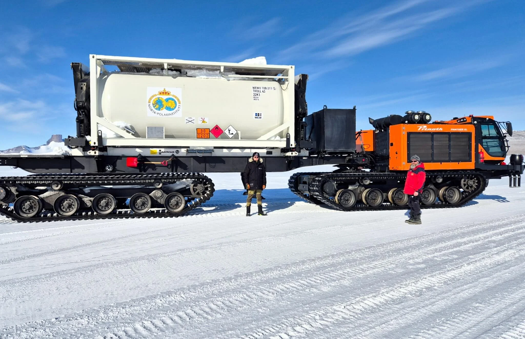 Two people standing in front of a Prinoth snow vehicle