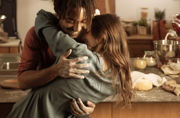 A young couple embraces warmly in a cozy kitchen while baking together, surrounded by dough, flour and fresh bread ingredients.