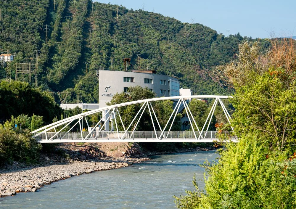 Reusch-Gebäude an einem Fluss mit einer weißen Fußgängerbrücke und einem bewaldeten Hang im Hintergrund.