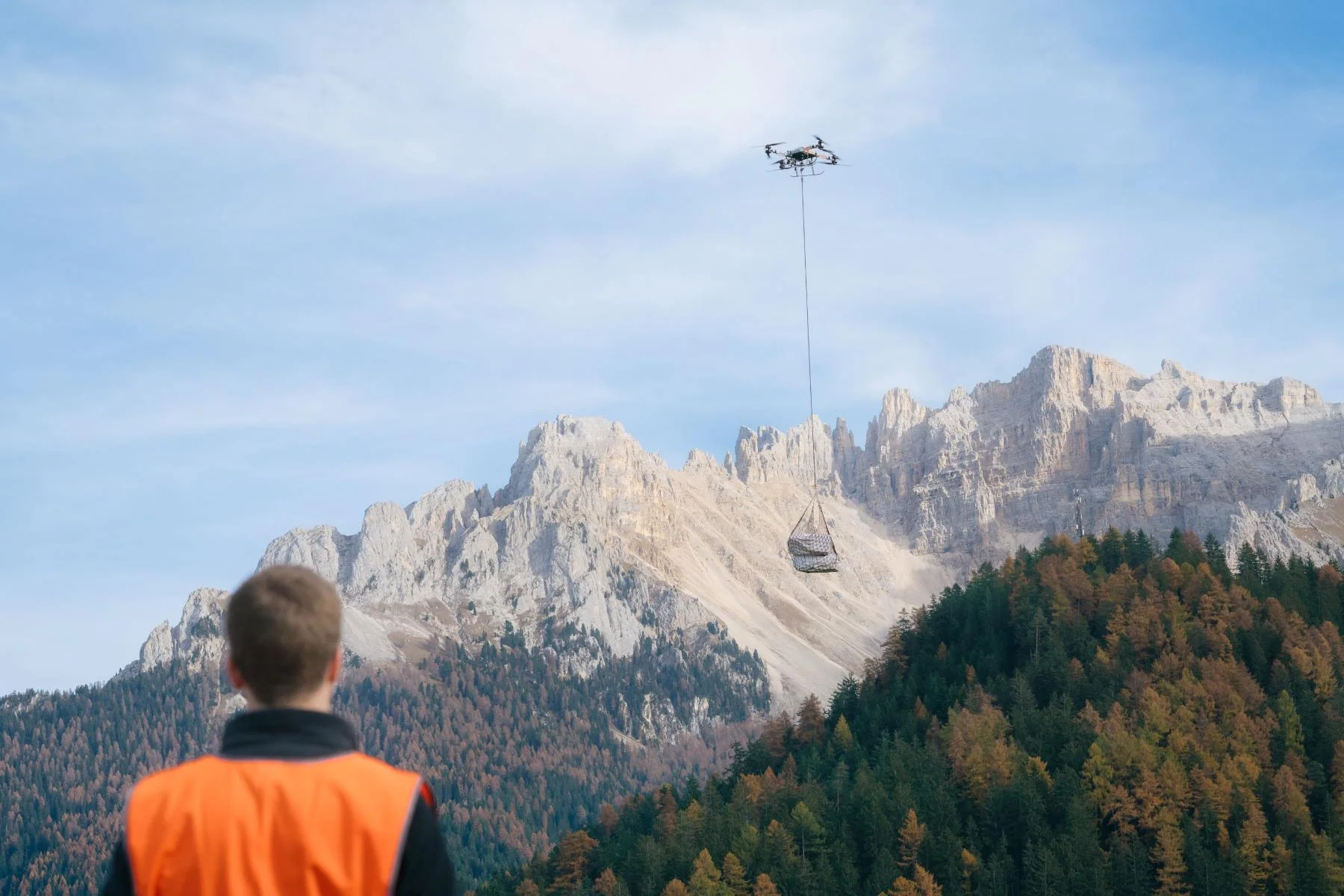 Cargo drone carrying an object, with the Dolomites in the background and a man in a safety vest in the foreground