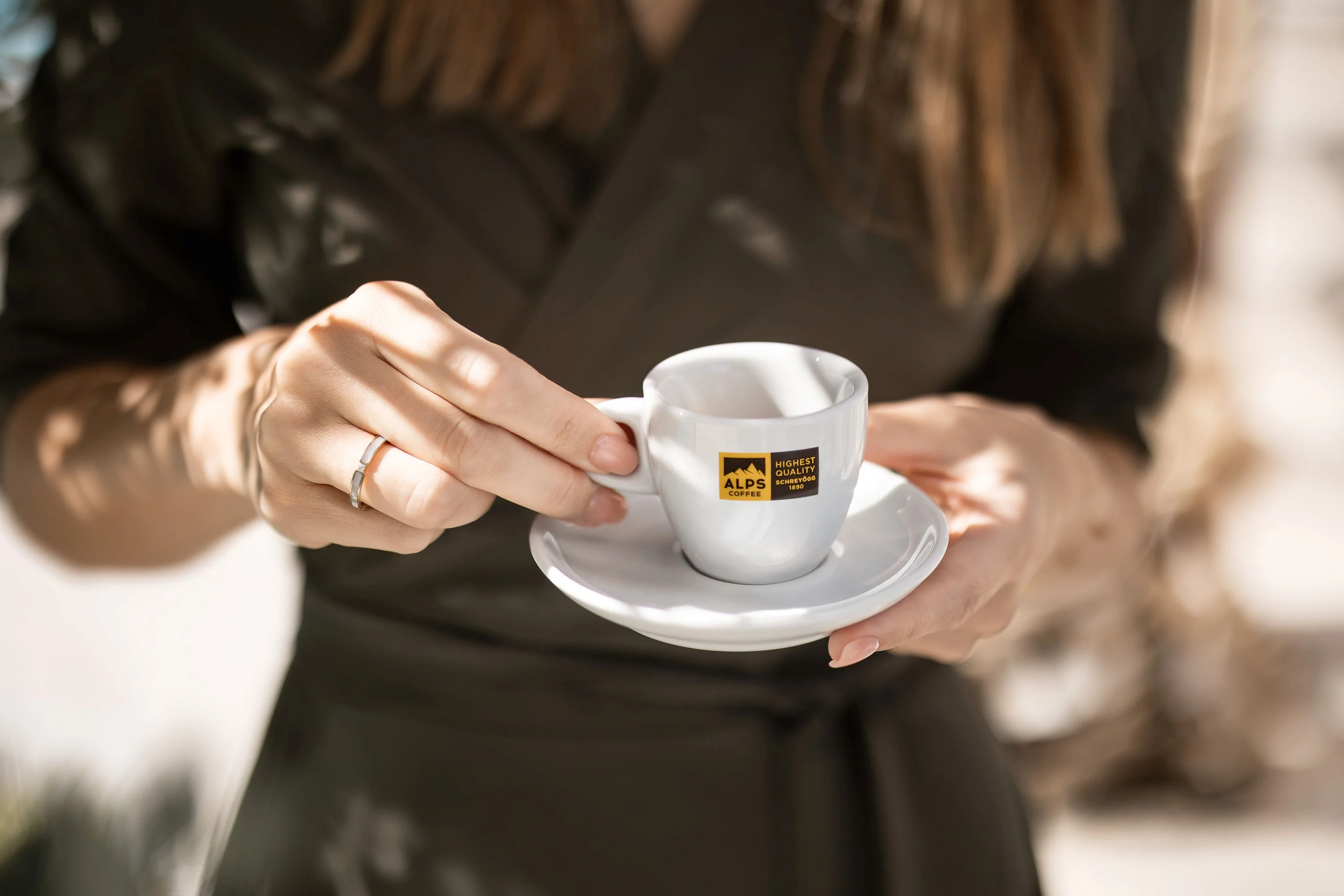 Nahaufnahme einer Person, die eine kleine Espressotasse mit Unterteller im warmen Sonnenlicht hält.