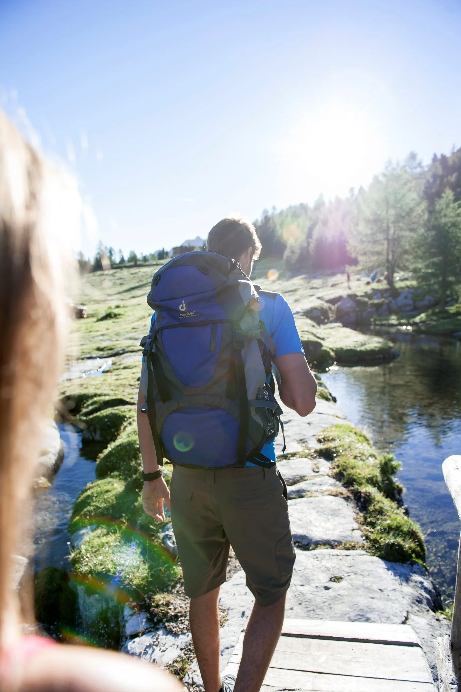 Wanderer mit Rucksack überquert einen Bach auf einem sonnigen alpinen Weg in Südtirol
