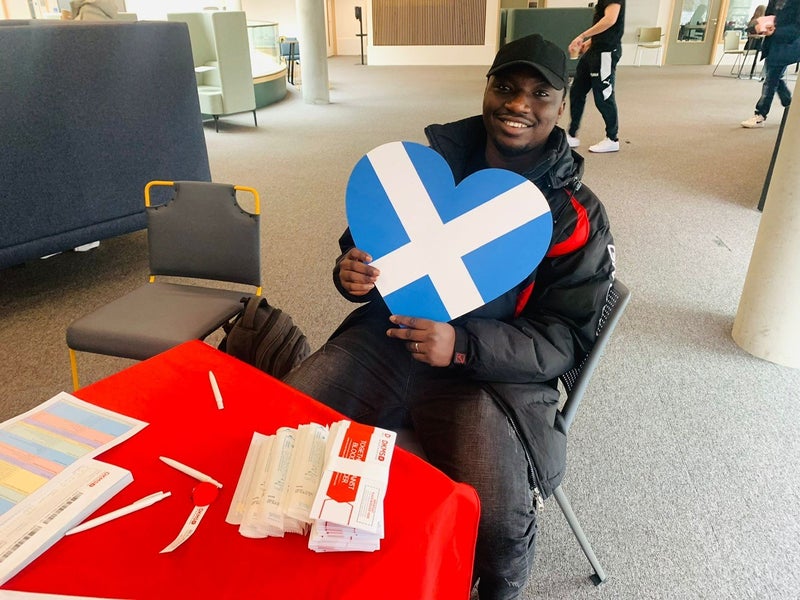 A DKMS volunteer at Stirling university smiling at holding  up a heart shaped Scottish flag 