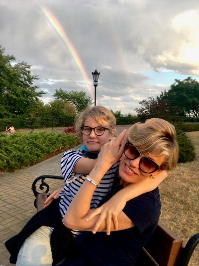 Amelia and her Mum sitting on a park bench. There is a double rainbow behind them in a grey sky. 