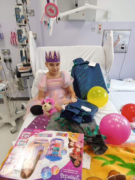Aashna sitting on her hospital bed wearing a paper crown, surroundd by colourful balloons and toys