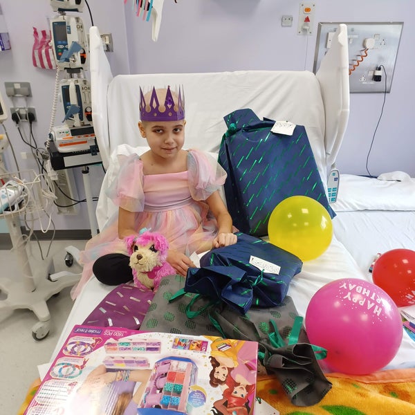 Aashna sitting on her hospital bed wearing a paper crown, surroundd by colourful balloons and toys