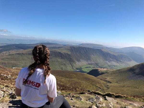 Image of a lady in a DKMS t-shirt sat on a mountain top