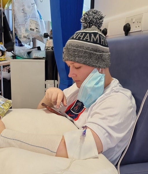 Kofi in hospital bed, reading his phone and wearing grey Tottenham Hotspur bobble hat