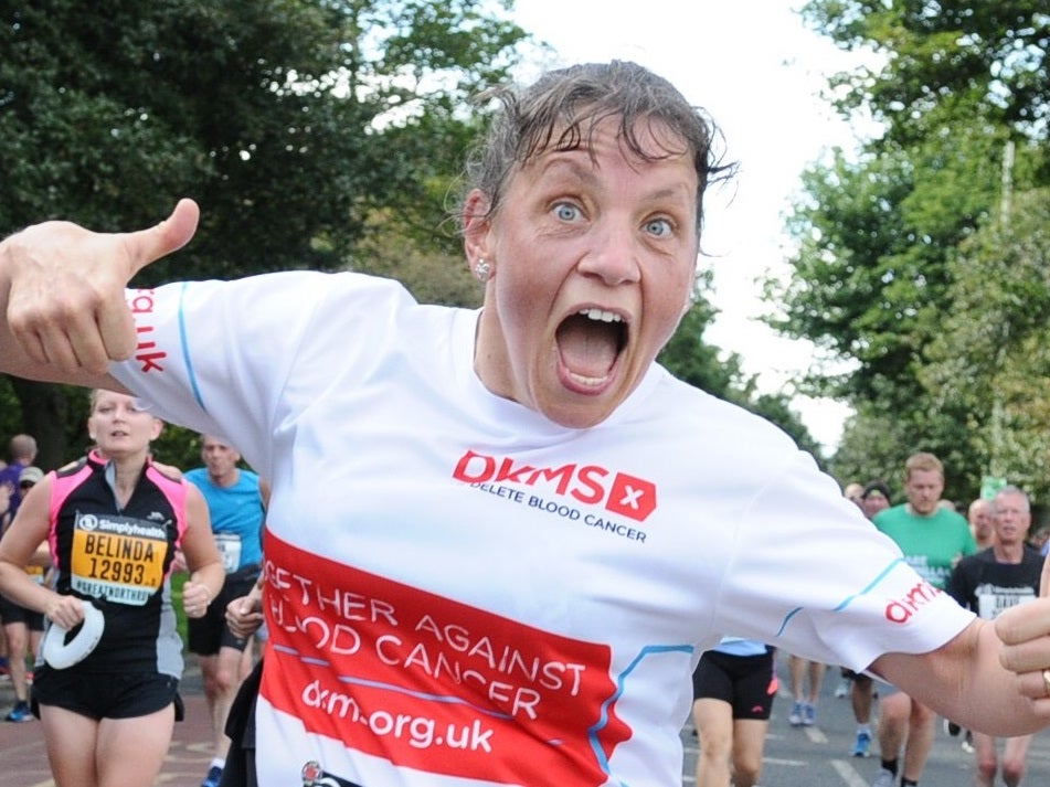 Woman wearing DKMS running top holds two thumbs up while running race, with other runners and supporters in the background. Woman wearing DKMS running top holds two thumbs up while running race, with other runners and supporters in the background.