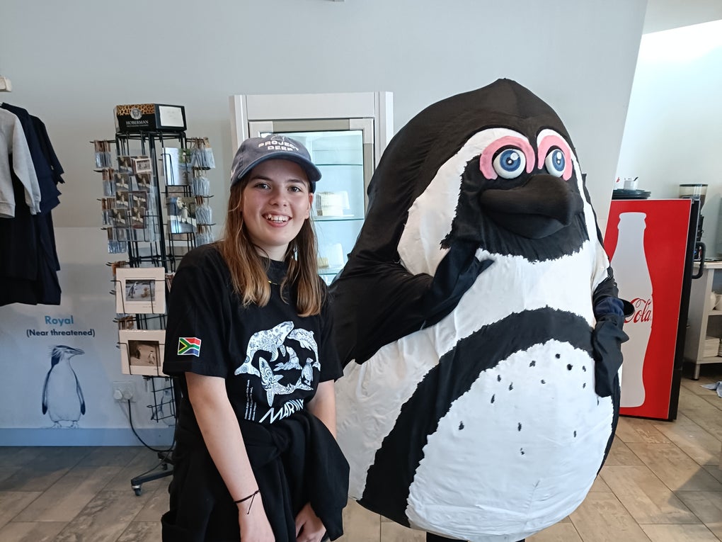 Alba wearing a graphic tee stood next to a penguin character Alba wearing a graphic tee stood next to a penguin character