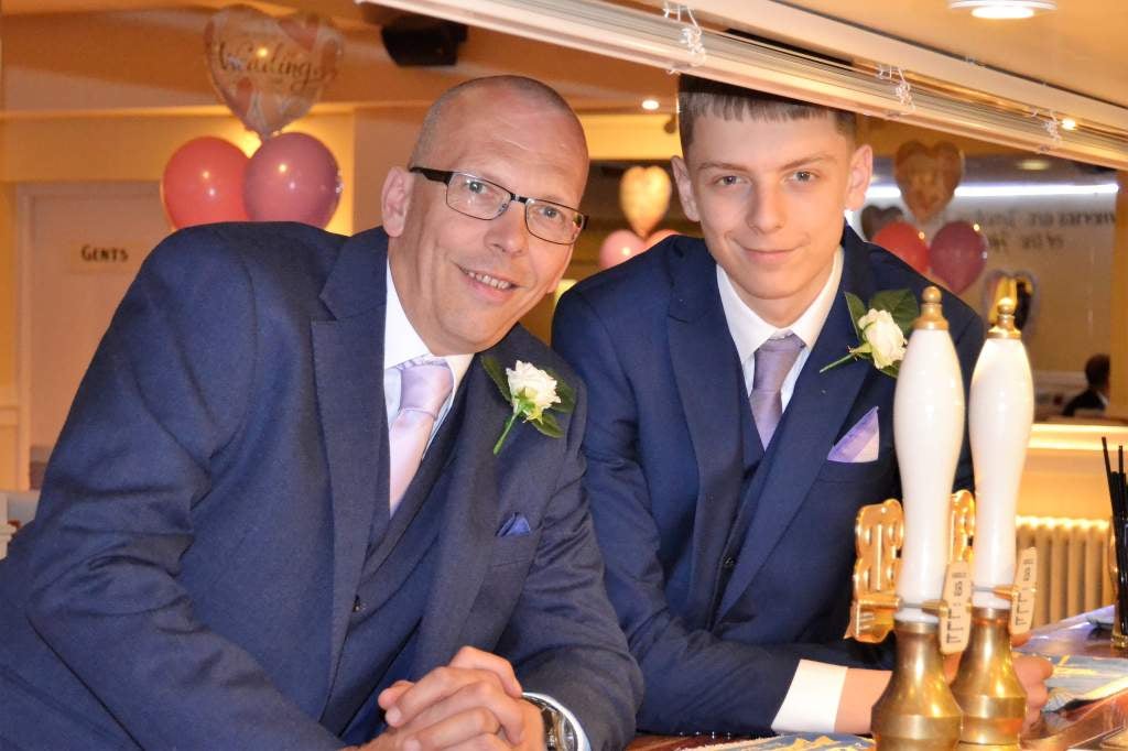 Luke and his Dad, Paul, stood at a bar celebrating his cousin's wedding. Both wearing blue suits and smiling into the camera. 