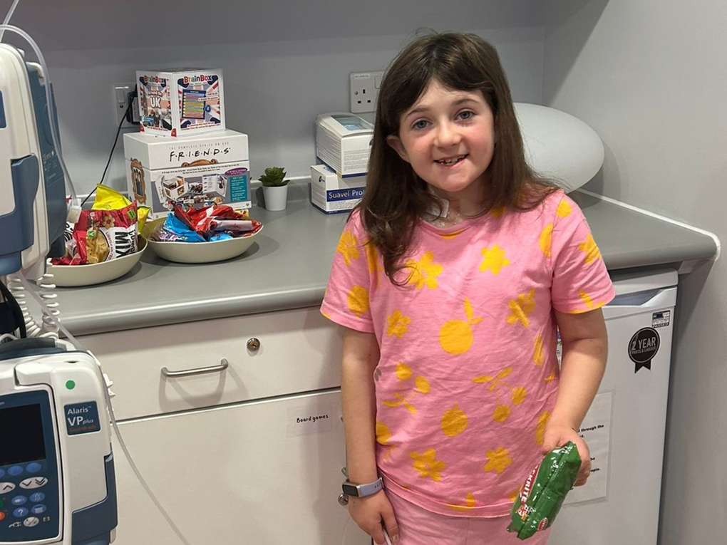 A little girl in hospital stands next to monitors, smiling and holding a bag of crisps. A little girl in hospital stands next to monitors, smiling and holding a bag of crisps.