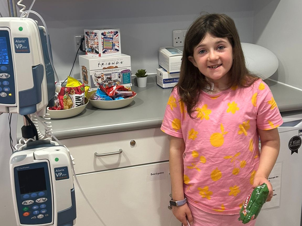 A little girl in hospital stands next to monitors, smiling and holding a bag of crisps.  A little girl in hospital stands next to monitors, smiling and holding a bag of crisps.