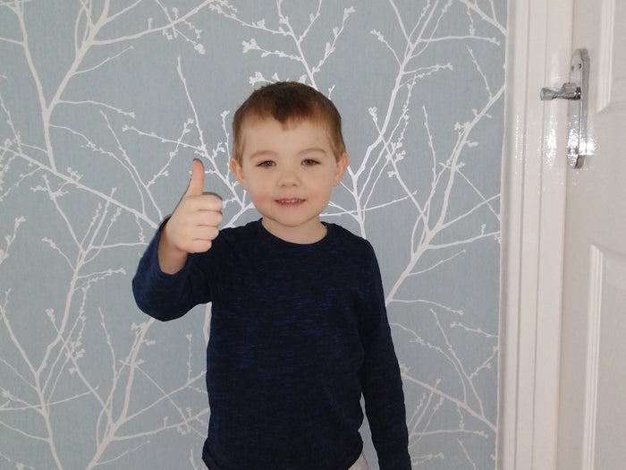 4 year old Emmett is standing with his right thumb up, he is smiling and looking at the camera, there is patterned wall paper in the background