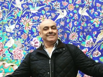 Shahin standing ini front of a blue patterned background, smiling and wearing a black coat. 