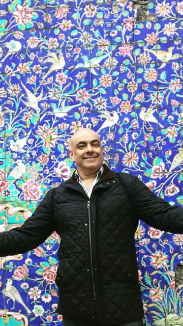 Shahin standing ini front of a blue patterned background, smiling and wearing a black coat. 