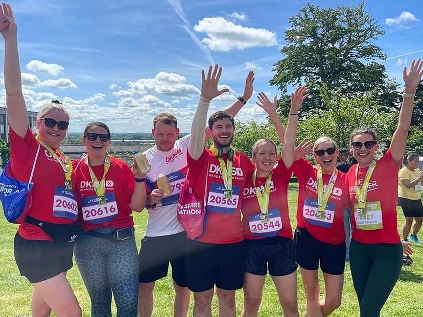 Group of runners stand on grass, holding their hands up in celebration, while wearing Red DKMS tops and race medals. Group of runners stand on grass, holding their hands up in celebration, while wearing Red DKMS tops and race medals.