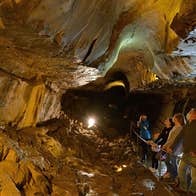 Five adults and one child standing on metal railed walkway looking on as a guide points upwards with a torch in a cavern with yellow lighting within the limestone cave