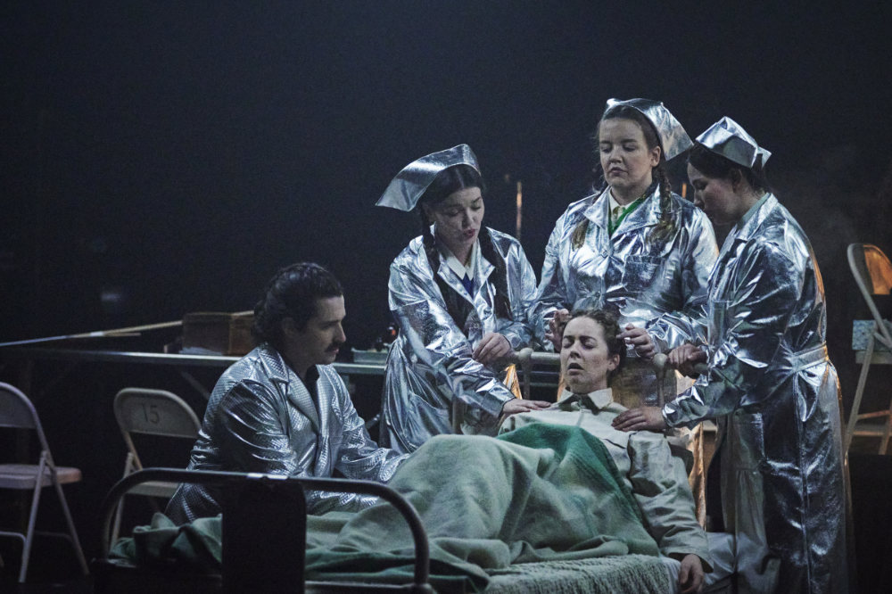 A scene from Elsewhere coming to Cork Opera House. A person is in a bed with 4 concerned people around the bed, all dressed in silver coloured coats and hats.