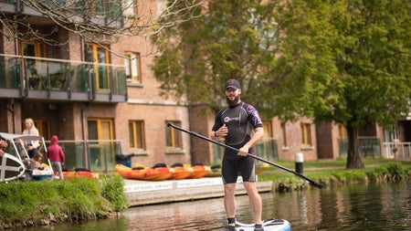Man in a wetsuit standing on a blue paddle board holding an oar on a canal