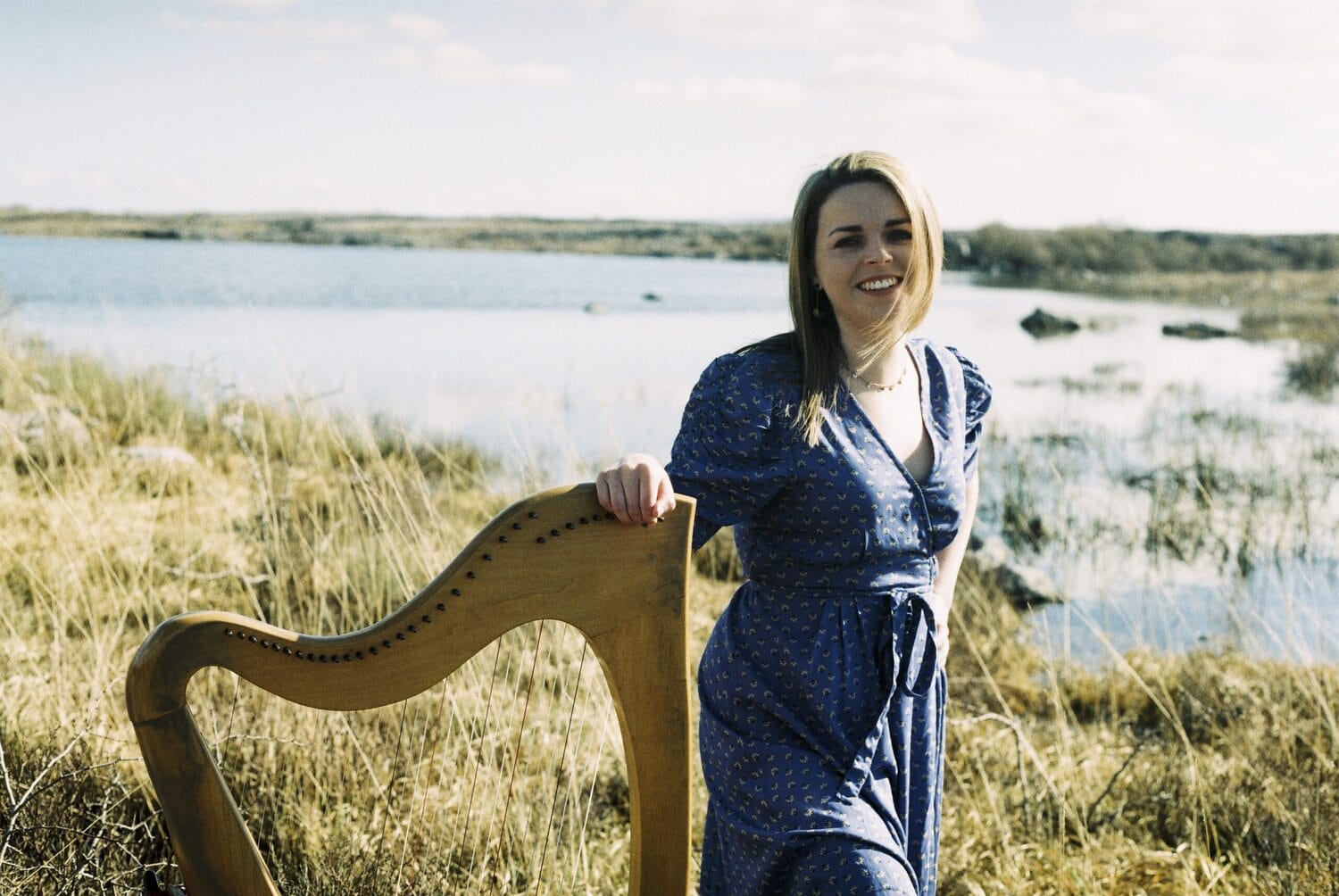 A woman is standing outside with a wooden harp beside her, a river and grass in the background.