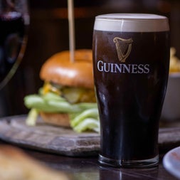 Pint of stout on a wooden counter with a blurred image of a burger behind it