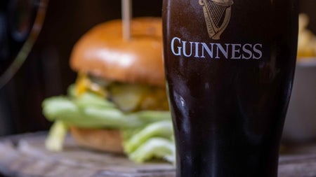 Pint of stout on a wooden counter with a blurred image of a burger behind it