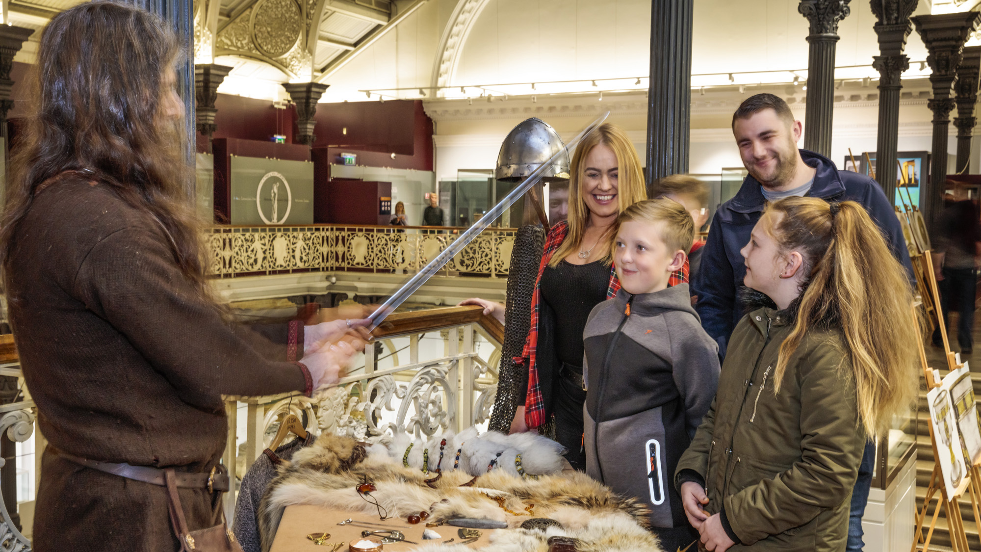 A family are standing at a table containing furs and other small artefacts with a man in brown costume holding up a sword.