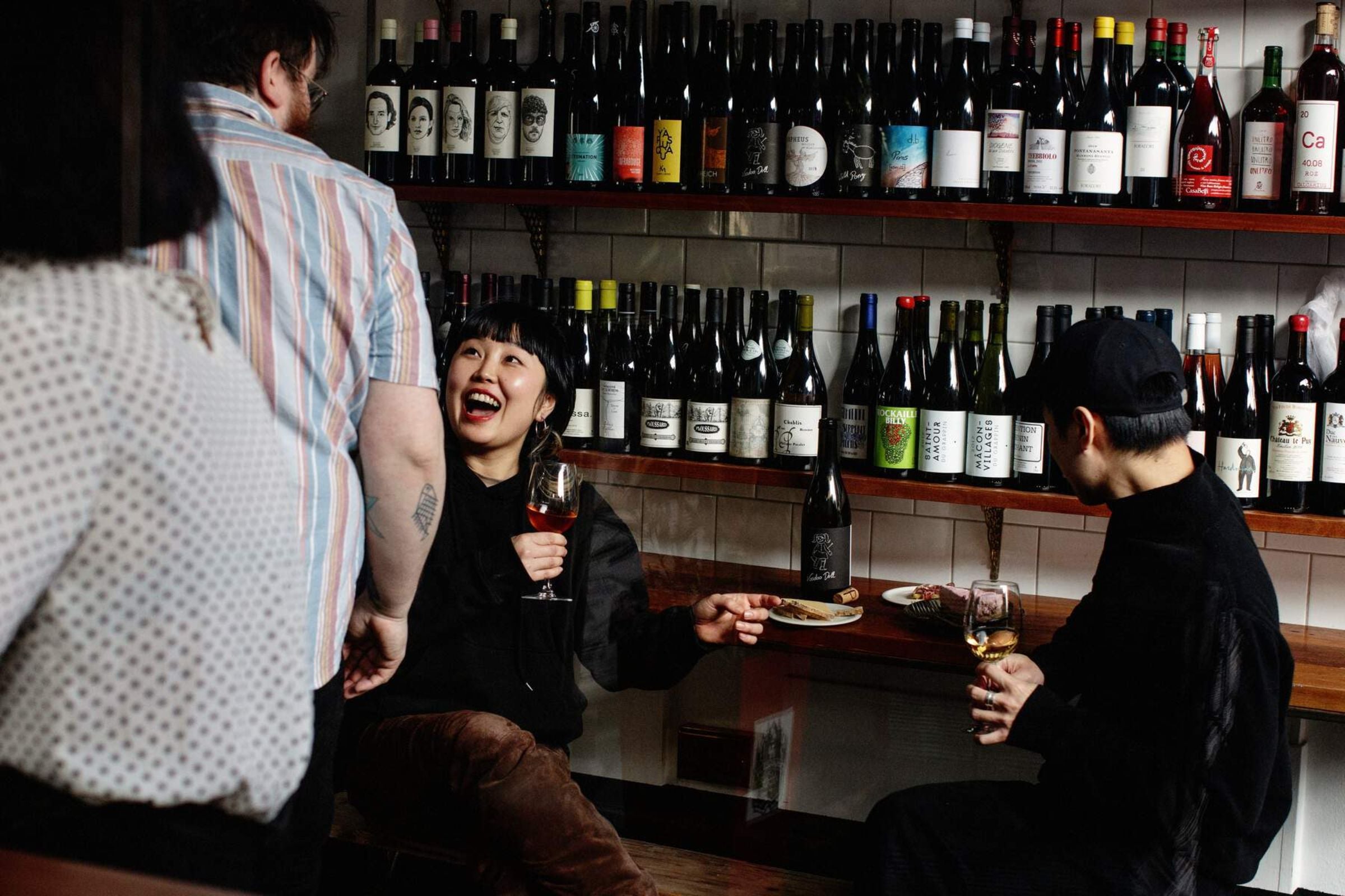 Woman holding a glass of wine laughing at a man passing her. There is two shelves of different wine bottles behind her on display.