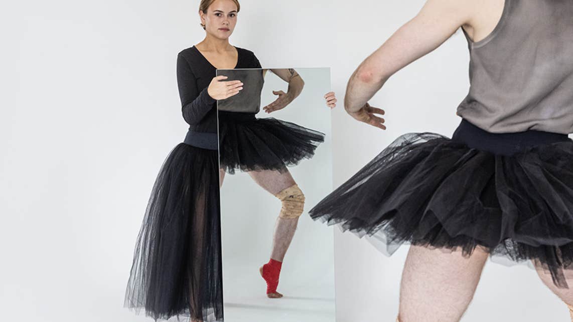 A person in black ballet outfit is holding a large mirror in front of them with reflection of another dancer standing in front, their face not visible.
