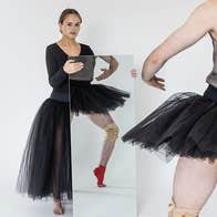 A person in black ballet outfit is holding a large mirror in front of them with reflection of another dancer standing in front, their face not visible.